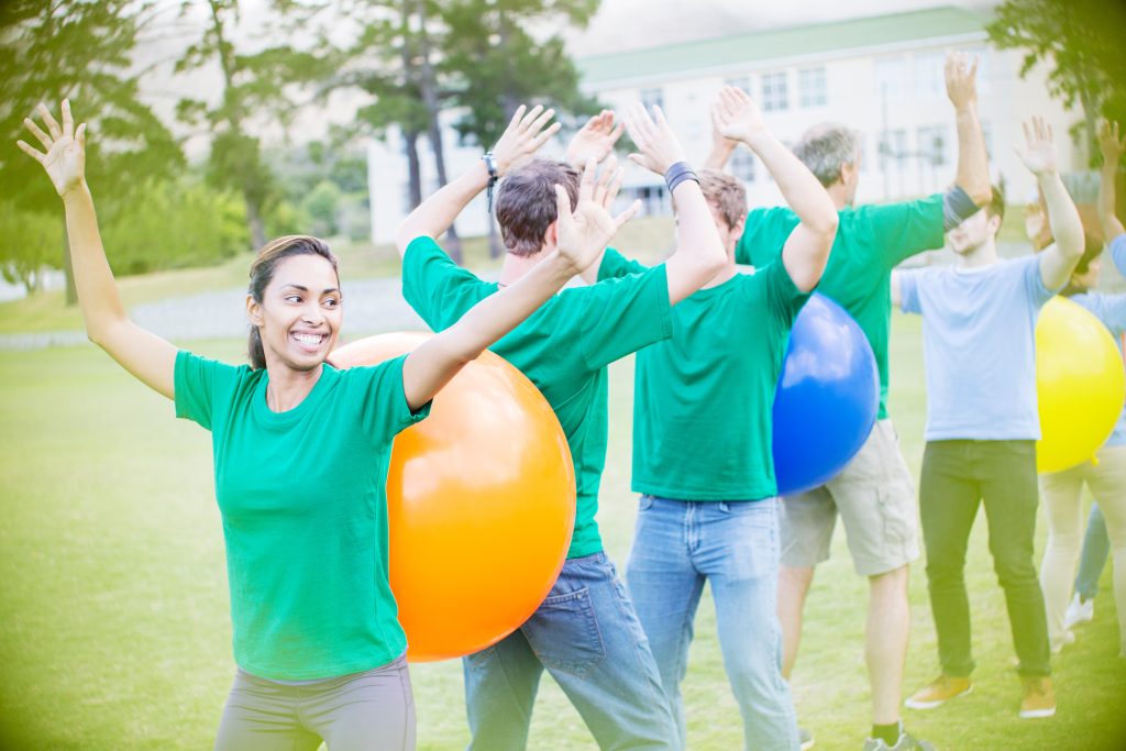 Groupe de jeunes adultes en pleine activité de team building en extérieur, formant une file avec des ballons colorés entre eux, souriants et levant les bras dans une ambiance conviviale.