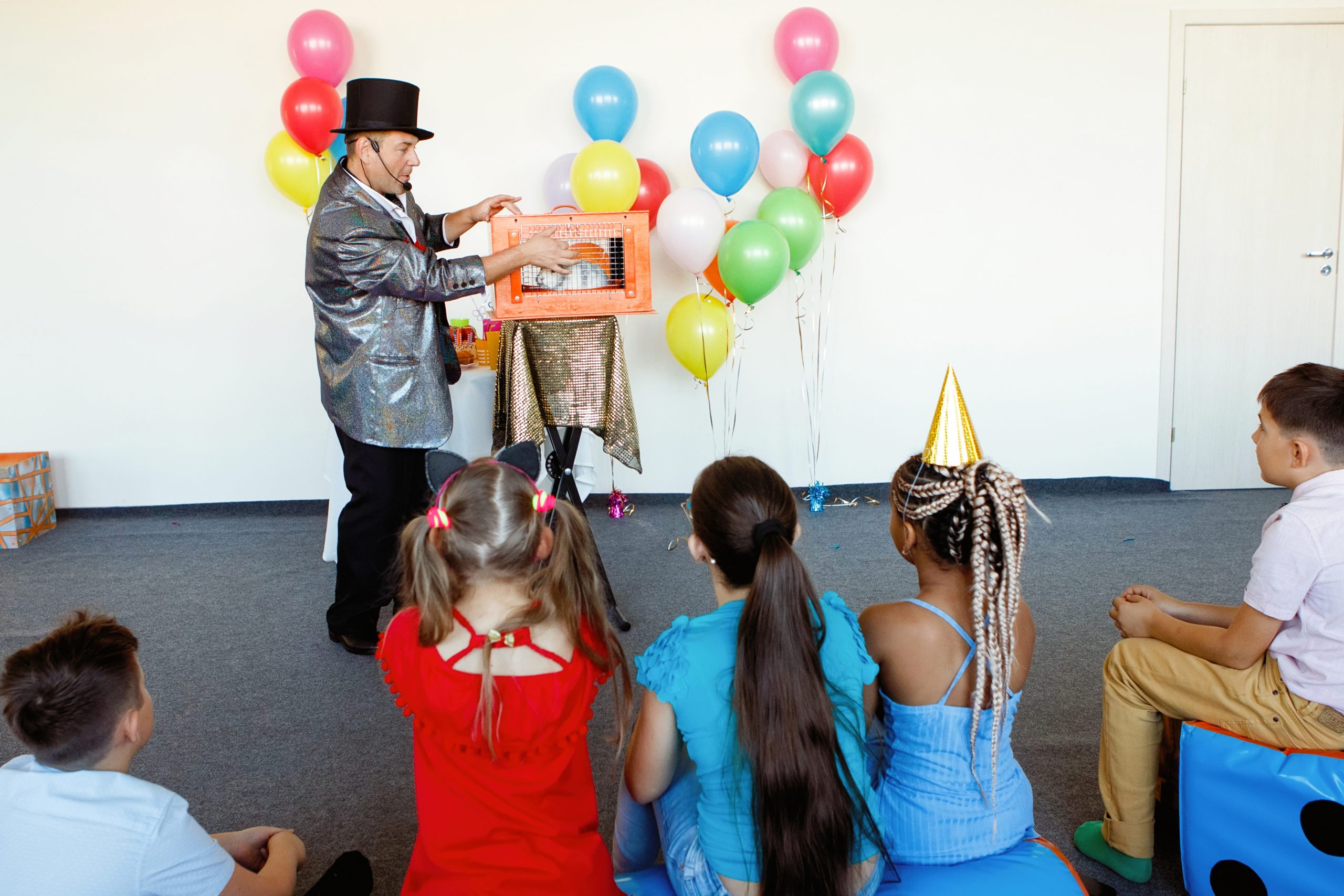 At a children's party, a magician, an illusionist shows tricks to a group of children in caps and with balloons.