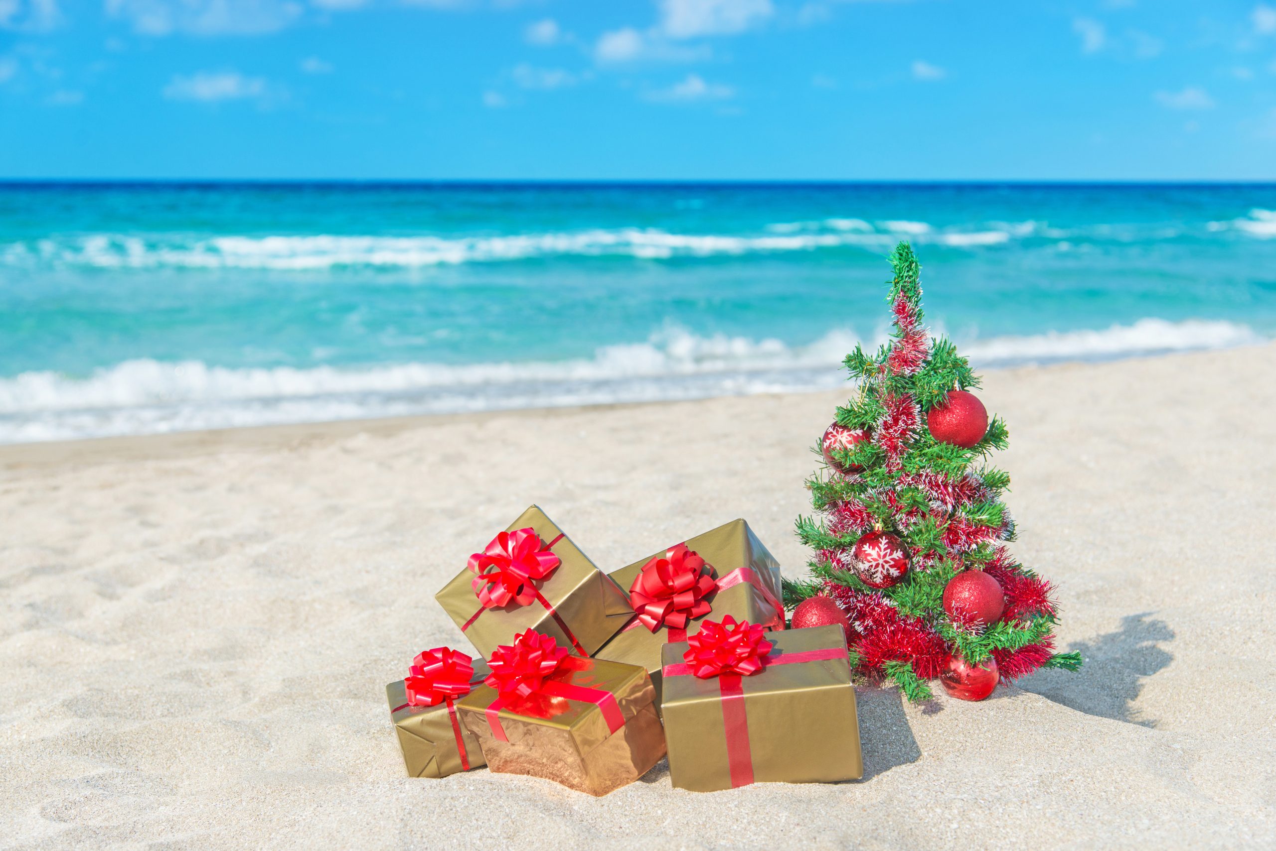 Christmas tree and golden gift with big red bow on the sea sandy beach. Christmas vacation concept.