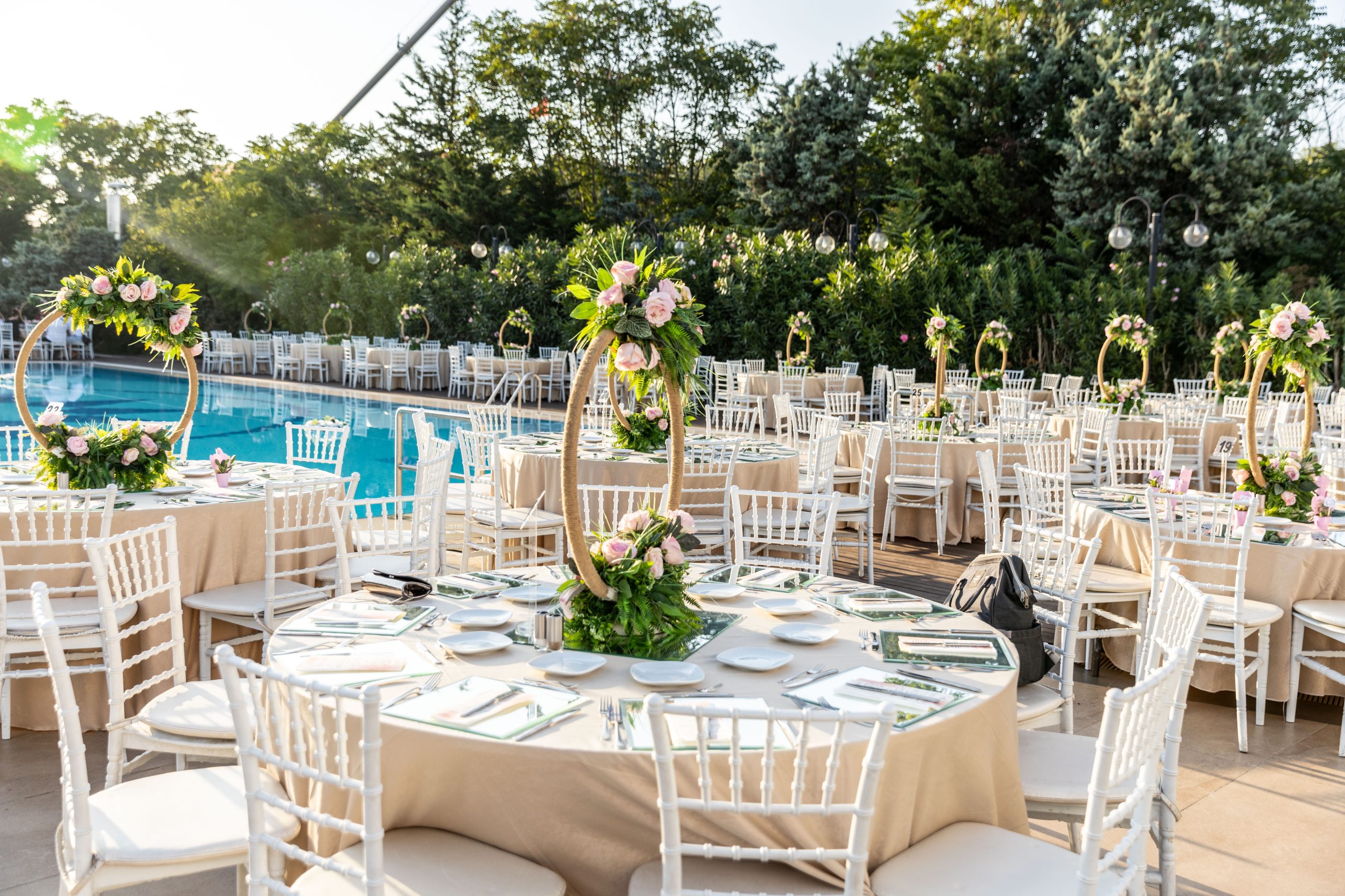 A stunning outdoor wedding reception with round tables, white chairs, and floral centerpieces. The tables are set with elegant glassware and silverware, creating a romantic and intimate atmosphere